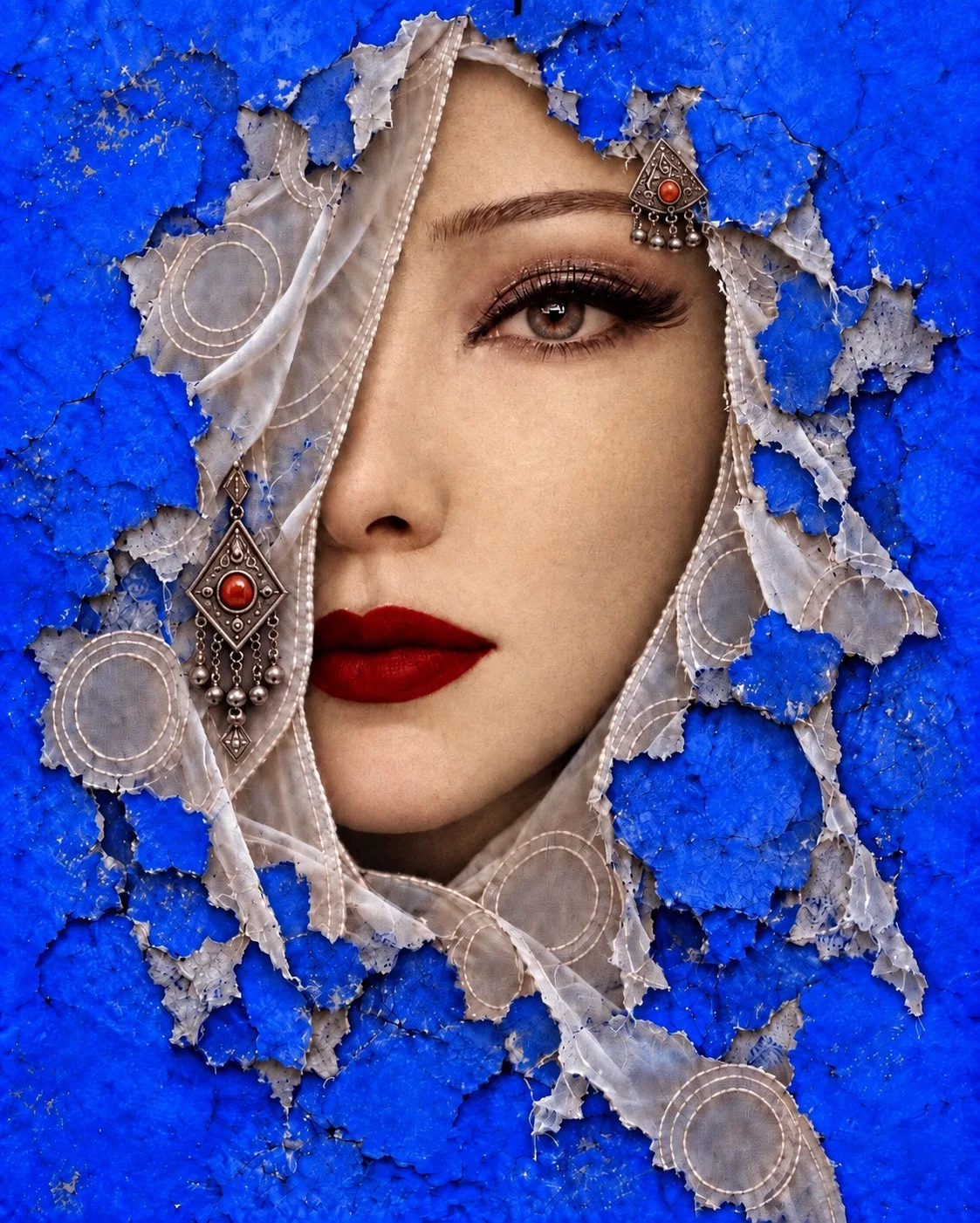 Visage encadré d’un voile de dentelle déchiré sur un fond bleu intense, bijou rouge.